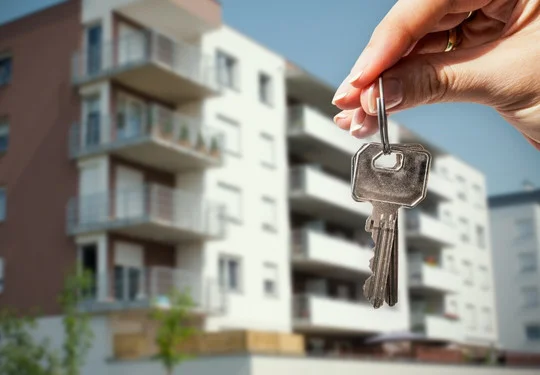 A hand holds a set of keys in the foreground with apartment buildings in the background